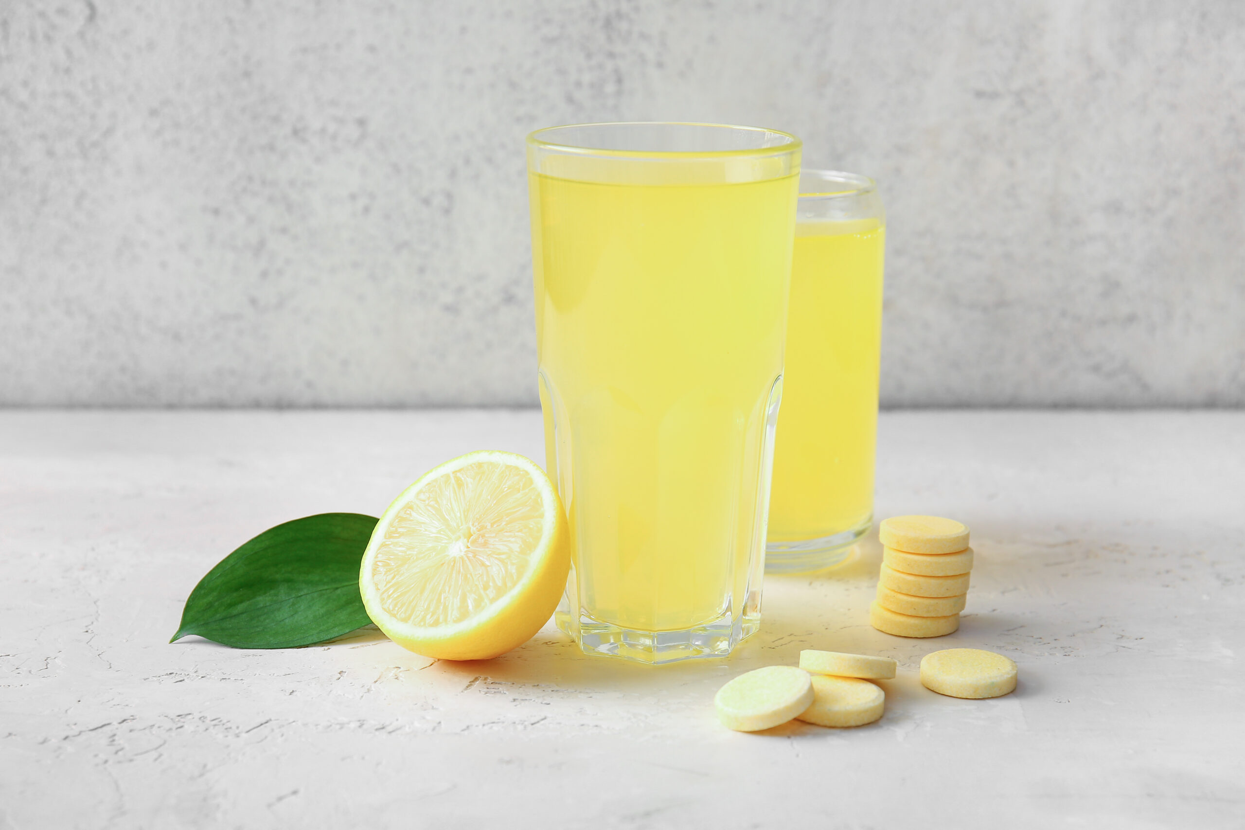 Glasses of vitamin C effervescent tablet dissolved in water and lemon on white grunge table